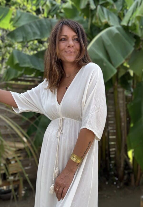 Robe longue fluide écrue – LENAE La robe LENAE écrue séduit par sa coupe longue, fluide et féminine. Elle offre un tomber naturel, parfait en toute occasion. Confectionnée en viscose légère, elle est agréable à porter même en plein été. Sa matière douce épouse les mouvements sans coller à la peau. Le décolleté en V se retrouve à l’avant comme à l’arrière, pour une touche féminine subtile. Sous la poitrine, un élastique fin souligne la taille sans comprimer. Un lien à nouer, orné de pompons, permet d’ajuster la robe avec charme et confort. Matière : viscose fluide Couleur : écrue Décolleté : devant et dos Manches : courtes Ceinture : lien à pompons Coupe : droite et évasée Taille : unique (36 à 40) Fabrication : Italie Associez-la à des sandales ou des espadrilles pour un look estival chic. Portez-la aussi en soirée avec une veste légère. ✨ Une robe pour briller au soleil, bouger librement, et se sentir belle en un clin d’œil. Attention : quantité très limitée !