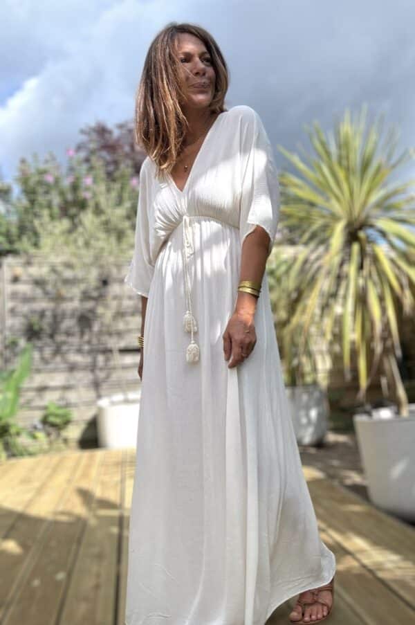Robe longue fluide écrue – LENAE La robe LENAE écrue séduit par sa coupe longue, fluide et féminine. Elle offre un tomber naturel, parfait en toute occasion. Confectionnée en viscose légère, elle est agréable à porter même en plein été. Sa matière douce épouse les mouvements sans coller à la peau. Le décolleté en V se retrouve à l’avant comme à l’arrière, pour une touche féminine subtile. Sous la poitrine, un élastique fin souligne la taille sans comprimer. Un lien à nouer, orné de pompons, permet d’ajuster la robe avec charme et confort. Matière : viscose fluide Couleur : écrue Décolleté : devant et dos Manches : courtes Ceinture : lien à pompons Coupe : droite et évasée Taille : unique (36 à 40) Fabrication : Italie Associez-la à des sandales ou des espadrilles pour un look estival chic. Portez-la aussi en soirée avec une veste légère. ✨ Une robe pour briller au soleil, bouger librement, et se sentir belle en un clin d’œil. Attention : quantité très limitée !